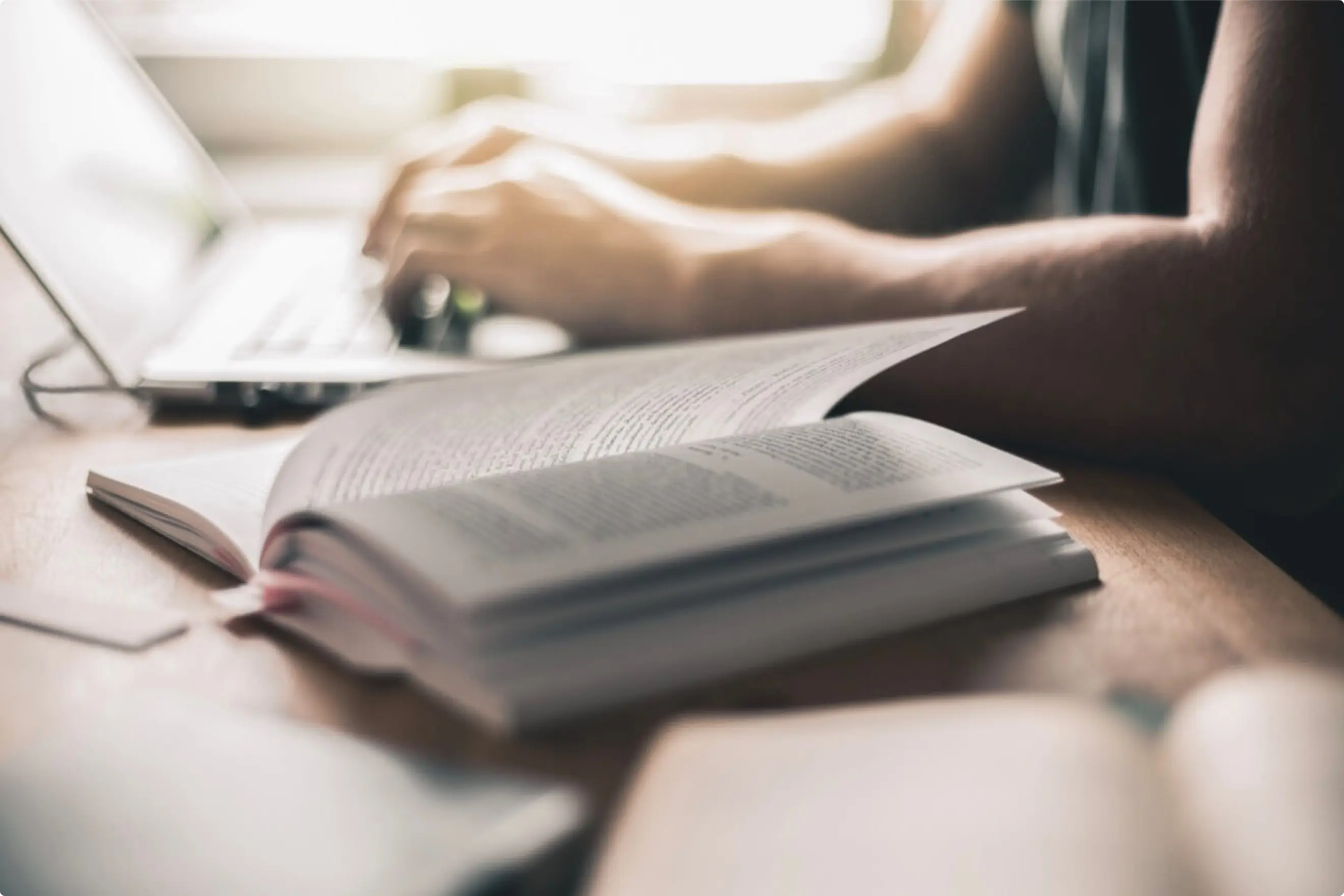 Im Vordergrund ein aufgeklapptes Buch und im Hintergrund sieht man zwei Hände und ein Notebook